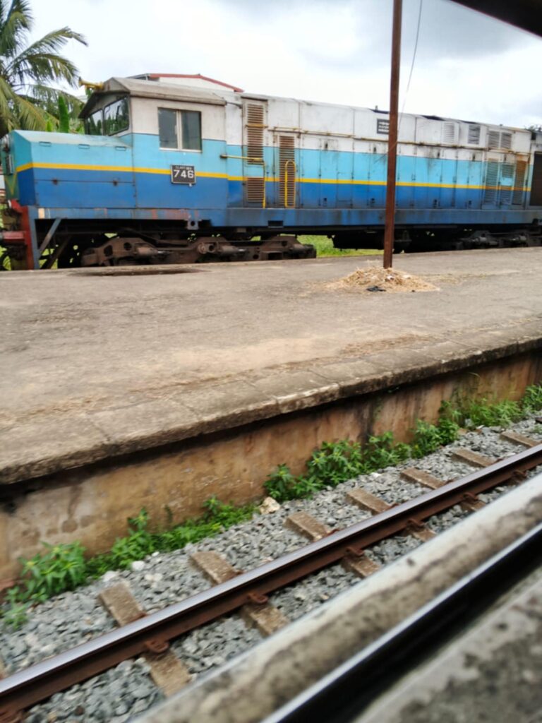Sri Lanka Railways diesel locomotive 746 at rural train station – side view captured from moving train window, featuring railway tracks and tropical background.