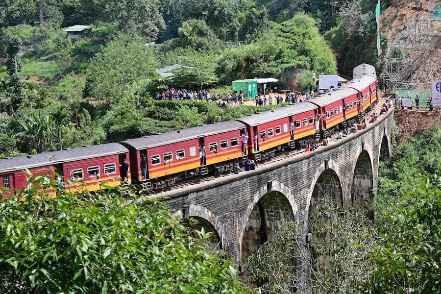 A red passenger train crossing the historic Nine Arch Bridge in Ella, Sri Lanka, surrounded by lush green jungle and entering a tunnel in the hillside.