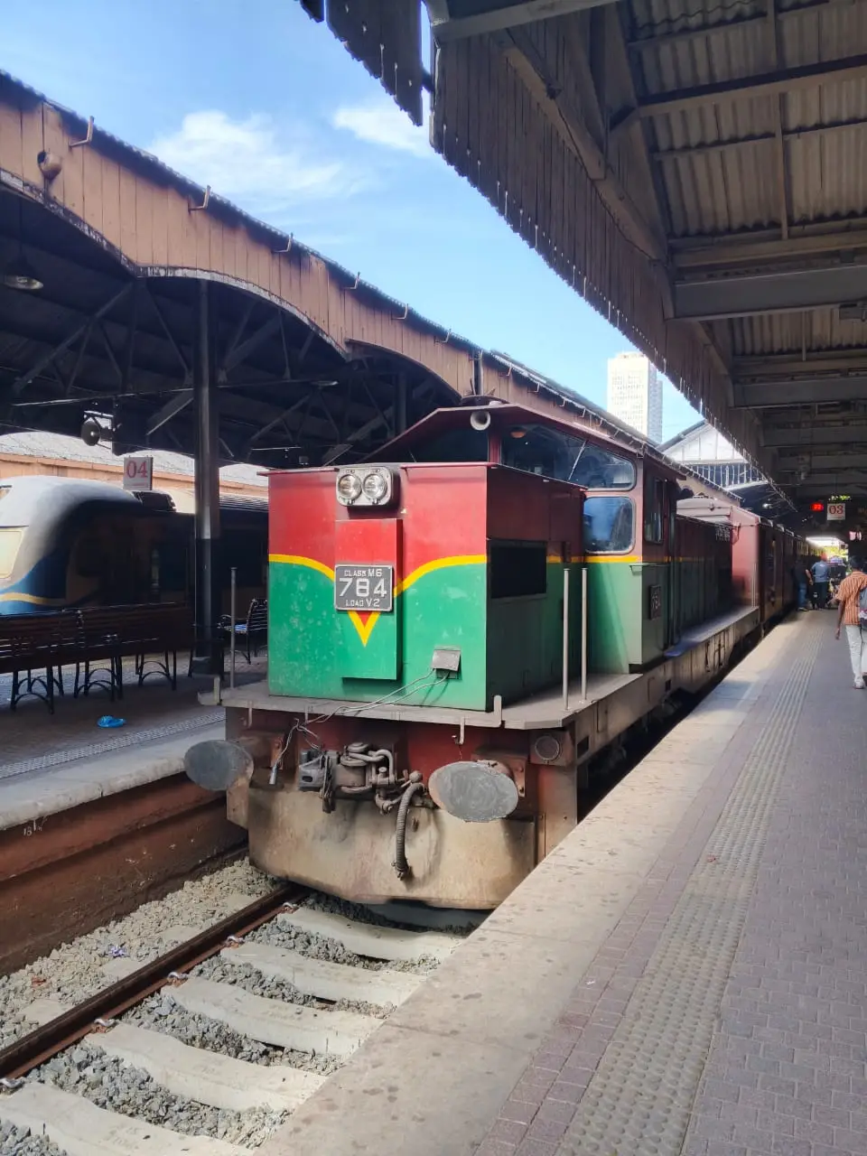Location: It appears to be a railway station, likely in Sri Lanka given the context of the current time and location (Colombo, Sri Lanka). This looks very much like Colombo Fort Railway Station. Locomotive: The locomotive is painted in a distinctive red and green livery, which is common for Sri Lanka Railways (SLR). Designation: The number plate clearly shows: "CLASS M6" and the unit number "784". Atmosphere: It's a sunny day, and the station has a covered platform area with an older, industrial feel.